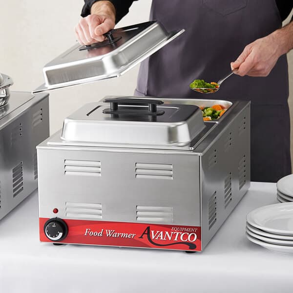 A person using a metal spoon to serve vegetables from a full size electric countertop food warmer.