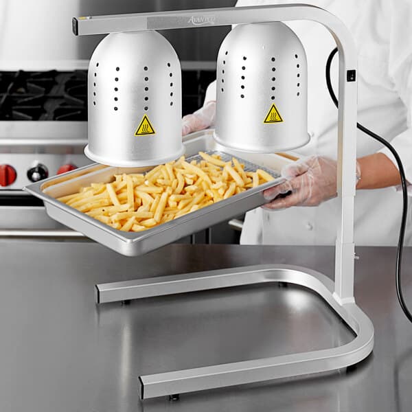 A person holding a tray of french fries under an Avantco countertop heat lamp.