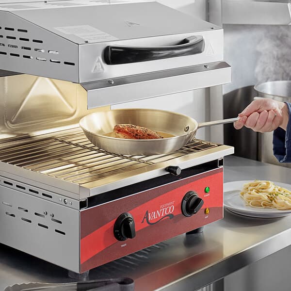 A person using an Avantco electric countertop salamander to cook meat in a pan.