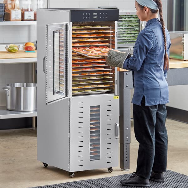 A woman standing in front of an Avantco stainless steel food dehydrator with glass doors.