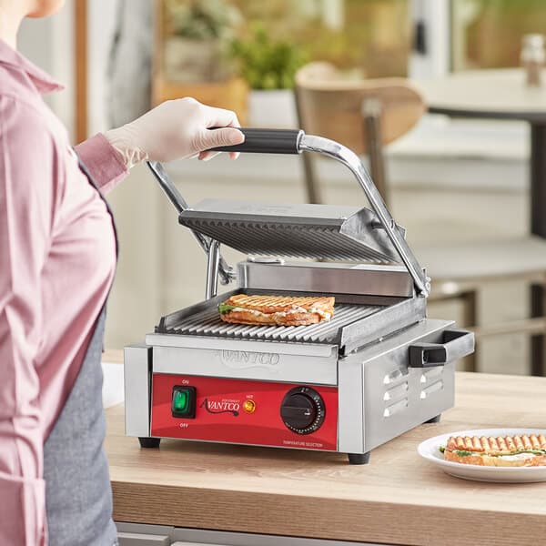 A woman using an Avantco Commercial Panini Grill to cook sandwiches on a table in a professional kitchen.