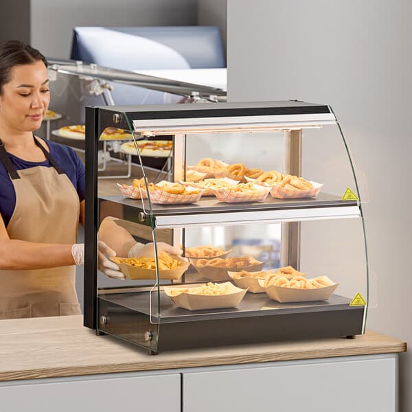 A woman wearing an apron standing behind a countertop display case with food in it.