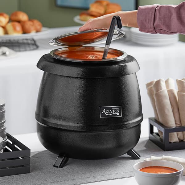 A woman pouring orange soup from a bowl into an Avantco black soup kettle.