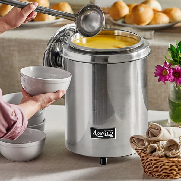 A woman holding a white bowl of soup in front of a stainless steel Avantco soup warmer.