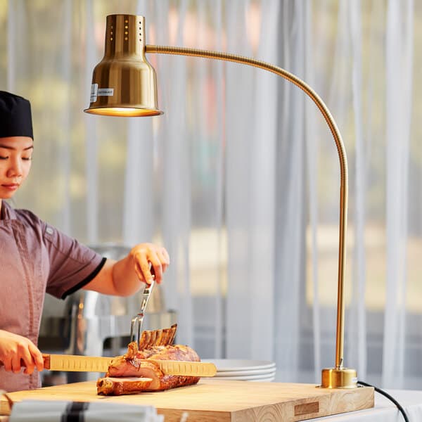 A woman cutting cooked meat on a cutting board with an Avantco gold flexible heat lamp above her.