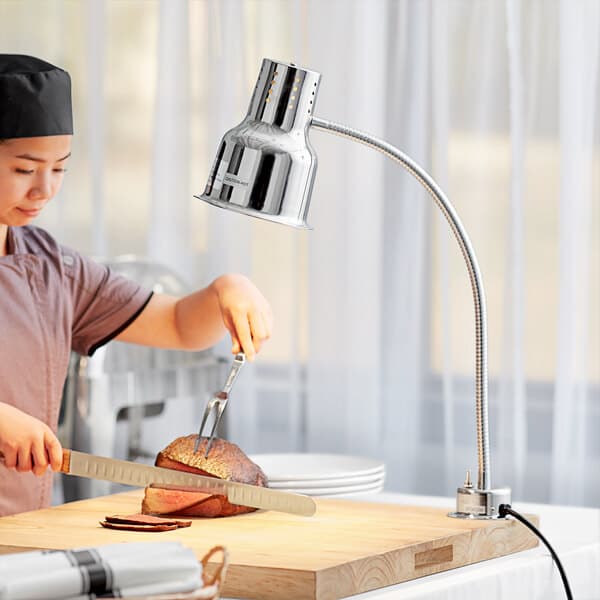 A woman using an Avantco countertop heat lamp to cut a piece of meat on a wooden surface.