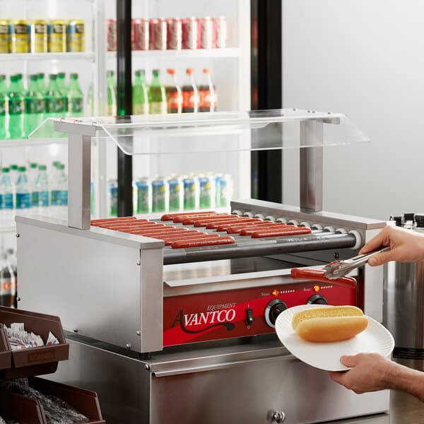 An Avantco Pass-Through Canopy sneeze guard on a hot dog roller grill with hot dogs on it.