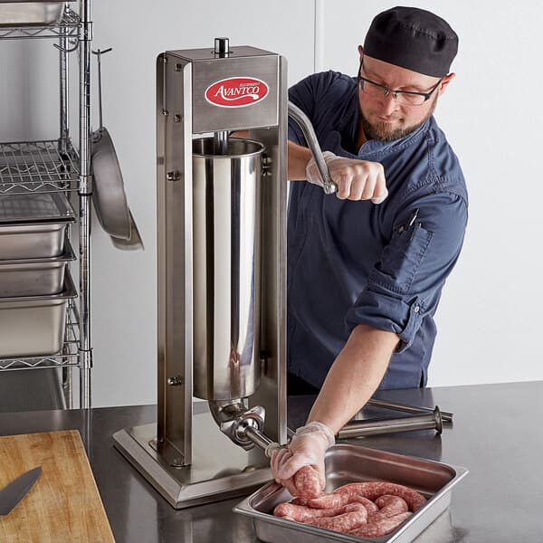 A man in a blue shirt and black hat using an Avantco stainless steel vertical manual sausage stuffer to make sausages.