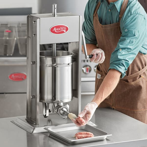 A man wearing an apron and gloves uses an Avantco stainless steel vertical manual sausage stuffer to make sausage.