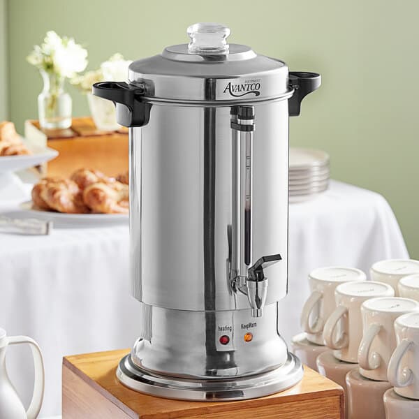 An Avantco stainless steel coffee urn on a wood table with white mugs.
