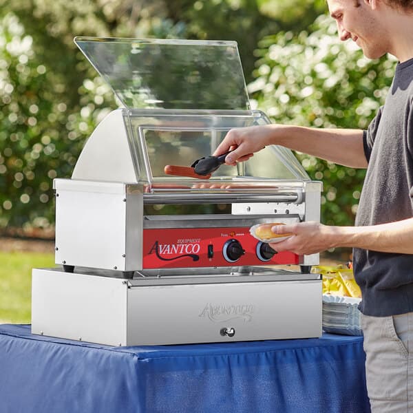 A man cooking hot dogs on an Avantco hot dog roller grill on a table outdoors.