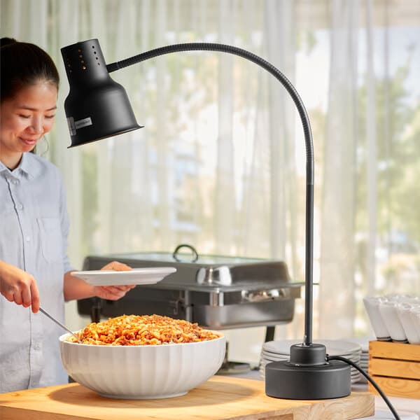 A woman holding a plate of food under a black Avantco countertop heat lamp.